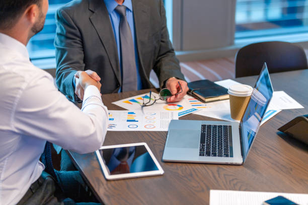 Two business professionals shaking hands over a table with charts and a laptop, symbolizing a business deal.