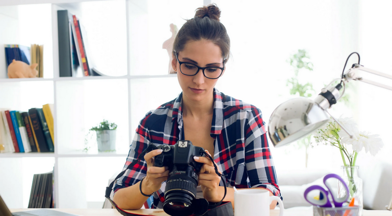 A person wearing glasses and a plaid shirt is inspecting a camera at a desk.