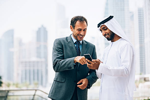 Two businessmen, one in a suit and the other in traditional attire, discuss something on a smartphone.